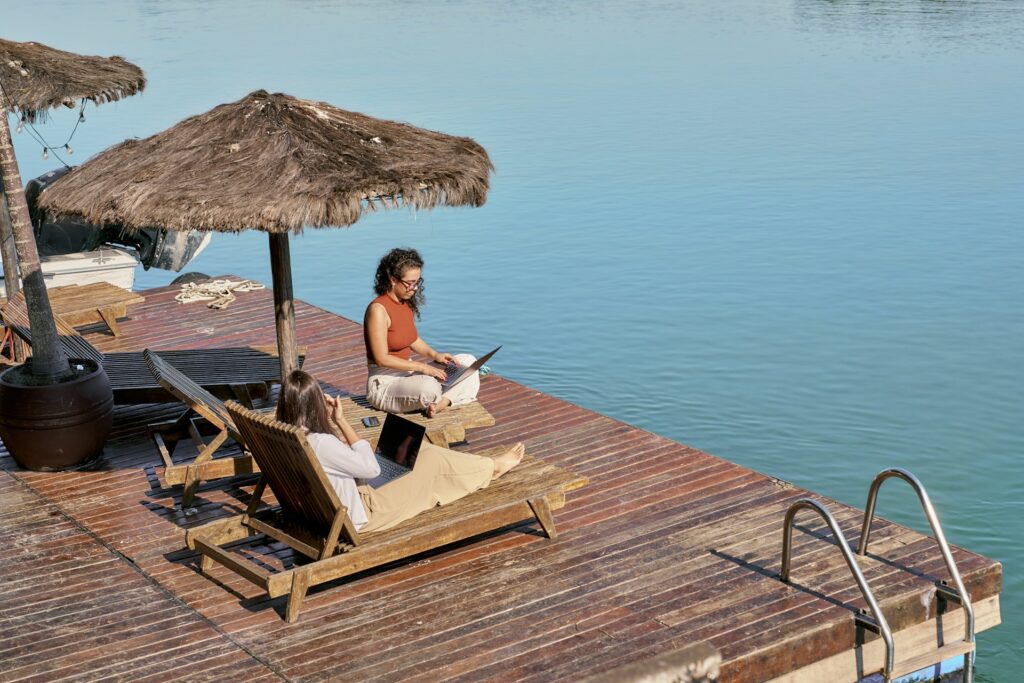 Two women working on their laptops on a dock ocean-side in a tropical locale, representing digital nomad workers