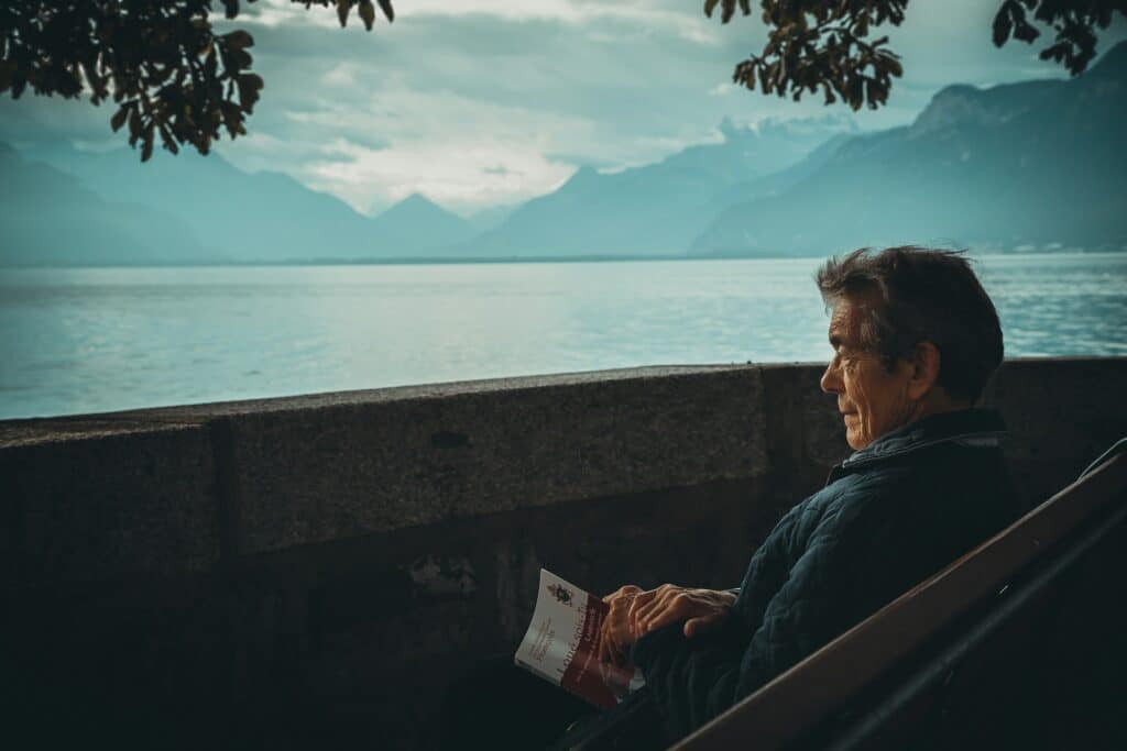 An elderly man sitting on a bench with a book looking out over water, representing enduring power of attorney.