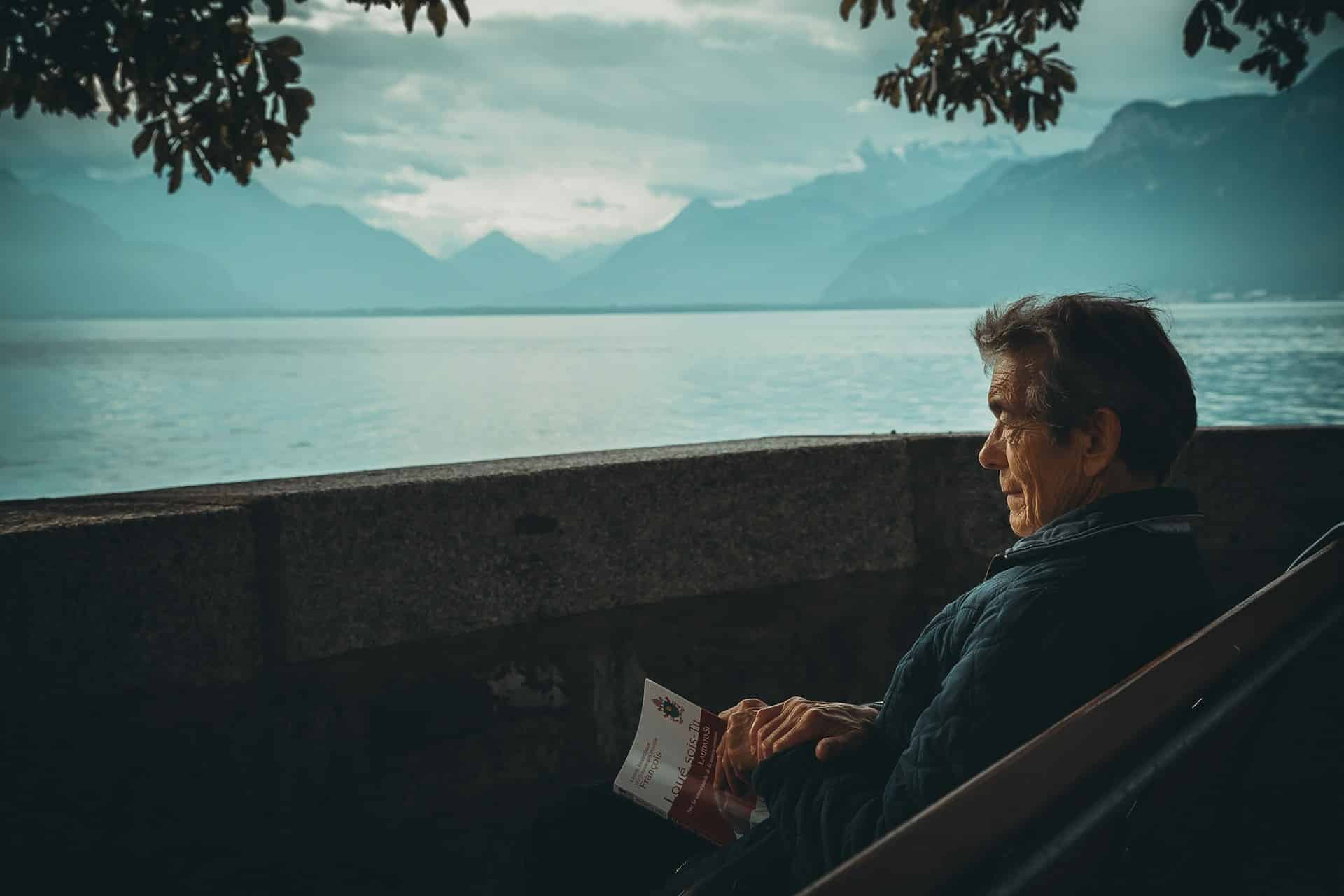 An elderly man sitting on a bench with a book looking out over water, representing enduring power of attorney.