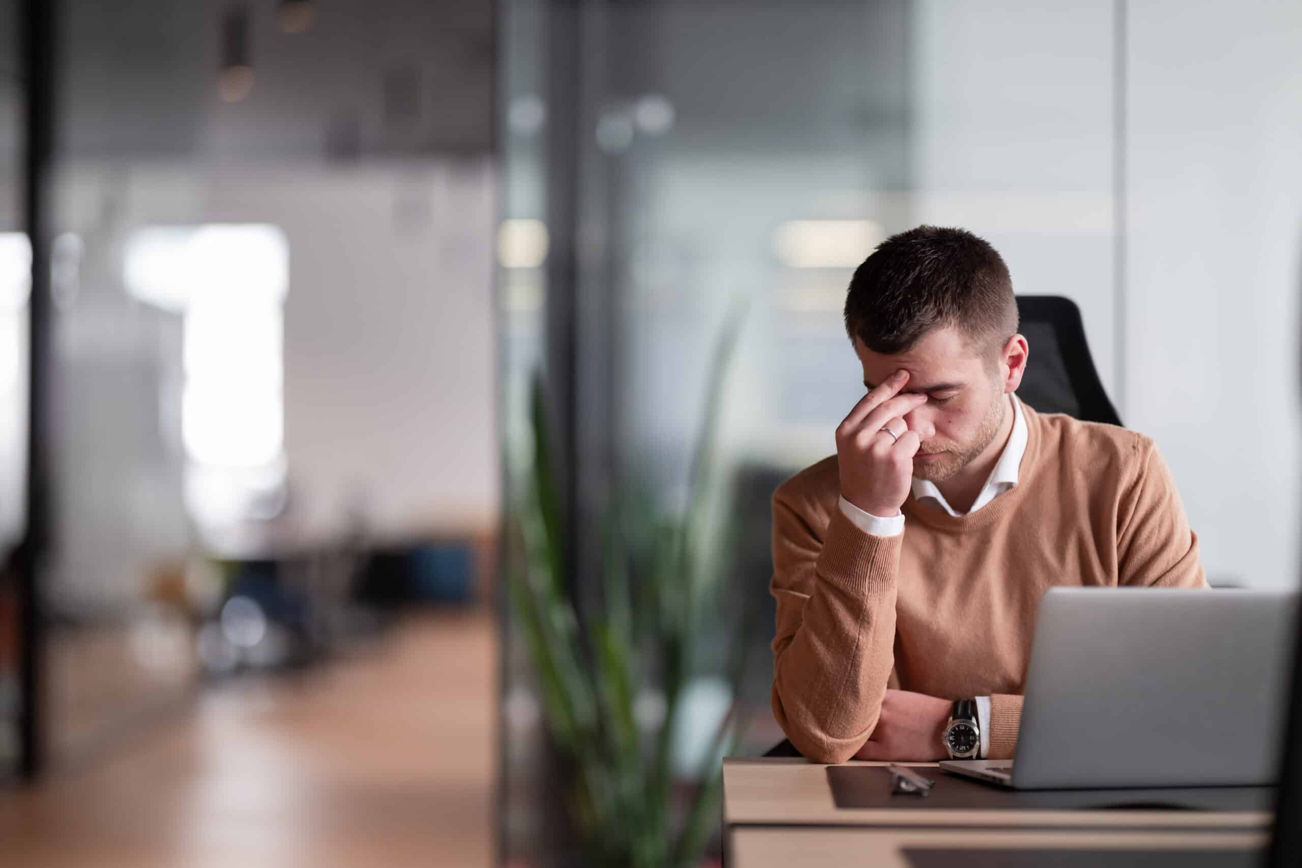An employee sitting at his desk, pinching the bridge of his nose, looking as if he's experiencing a headache or stress, representing Wrongful Dismissal law in Calgary, Alberta.