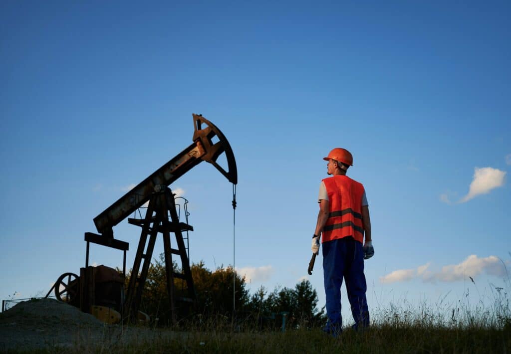 A worker in an orange hard hat and bright orange safety vest stands in a grassy field, looking towards a silhouetted oil pump jack against a clear blue sky at dusk.