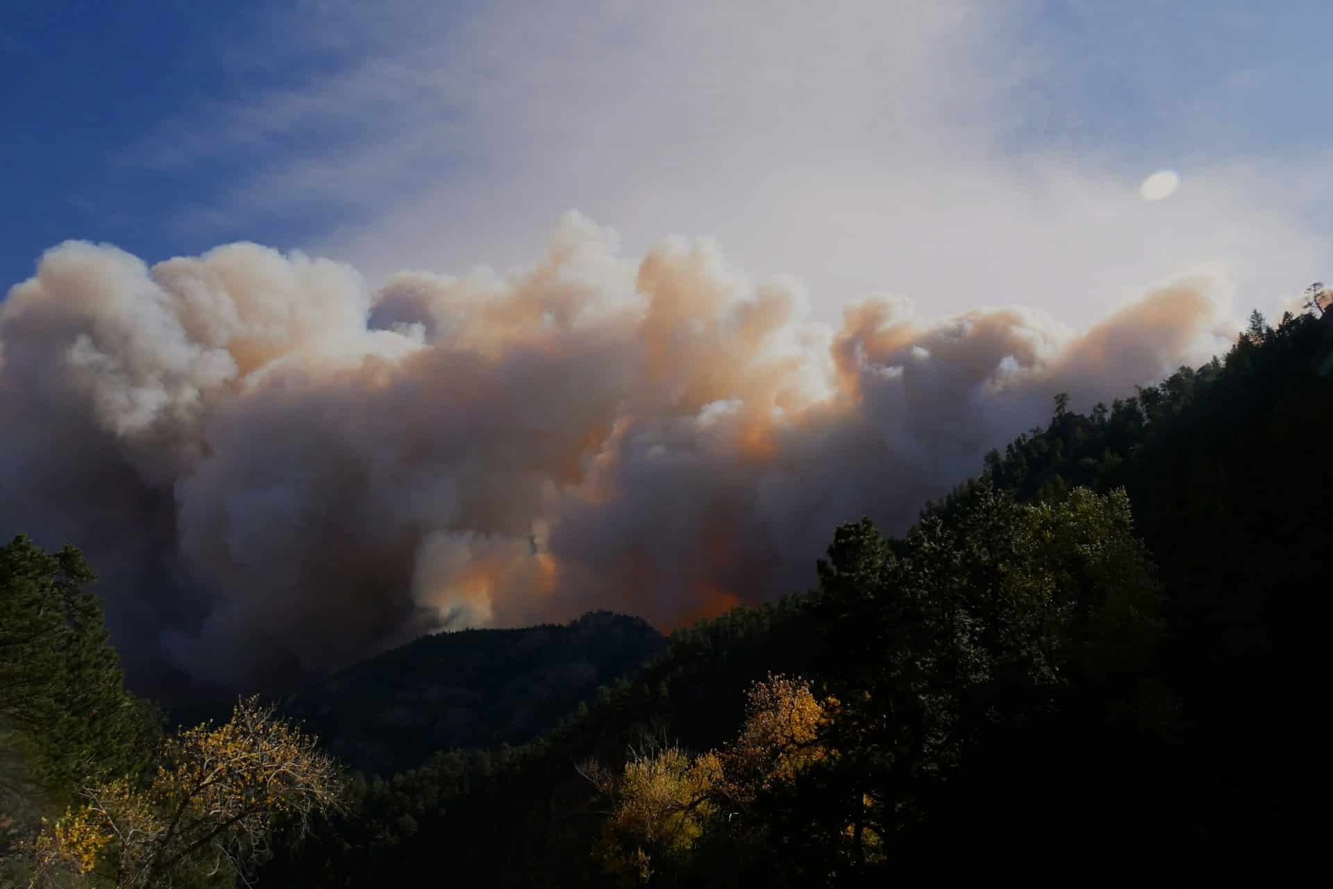 Billowing clouds of smoke from a wildfire blot out the blue sky and cast a shadow over tree-covered mountains, representing the type of natural disaster that necessitates force majeure clauses.