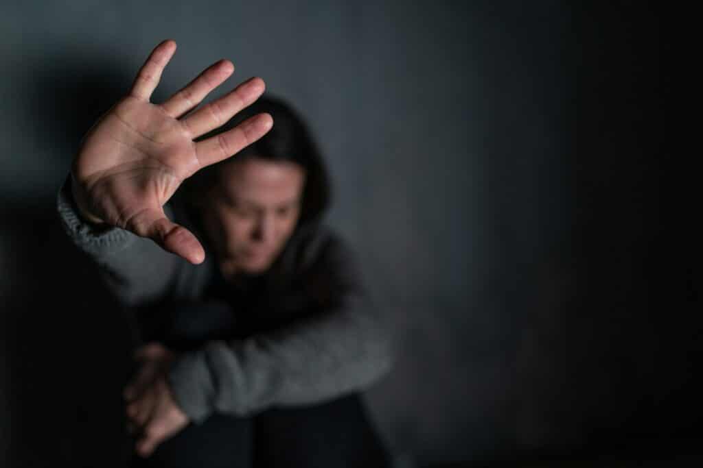 A visibly distressed woman is crouched on the floor against a wall with one arm wrapped around her knees, drawn to her chest, while the other is raised with her hand up and palm out, representing family violence and emergency protection orders in Alberta.