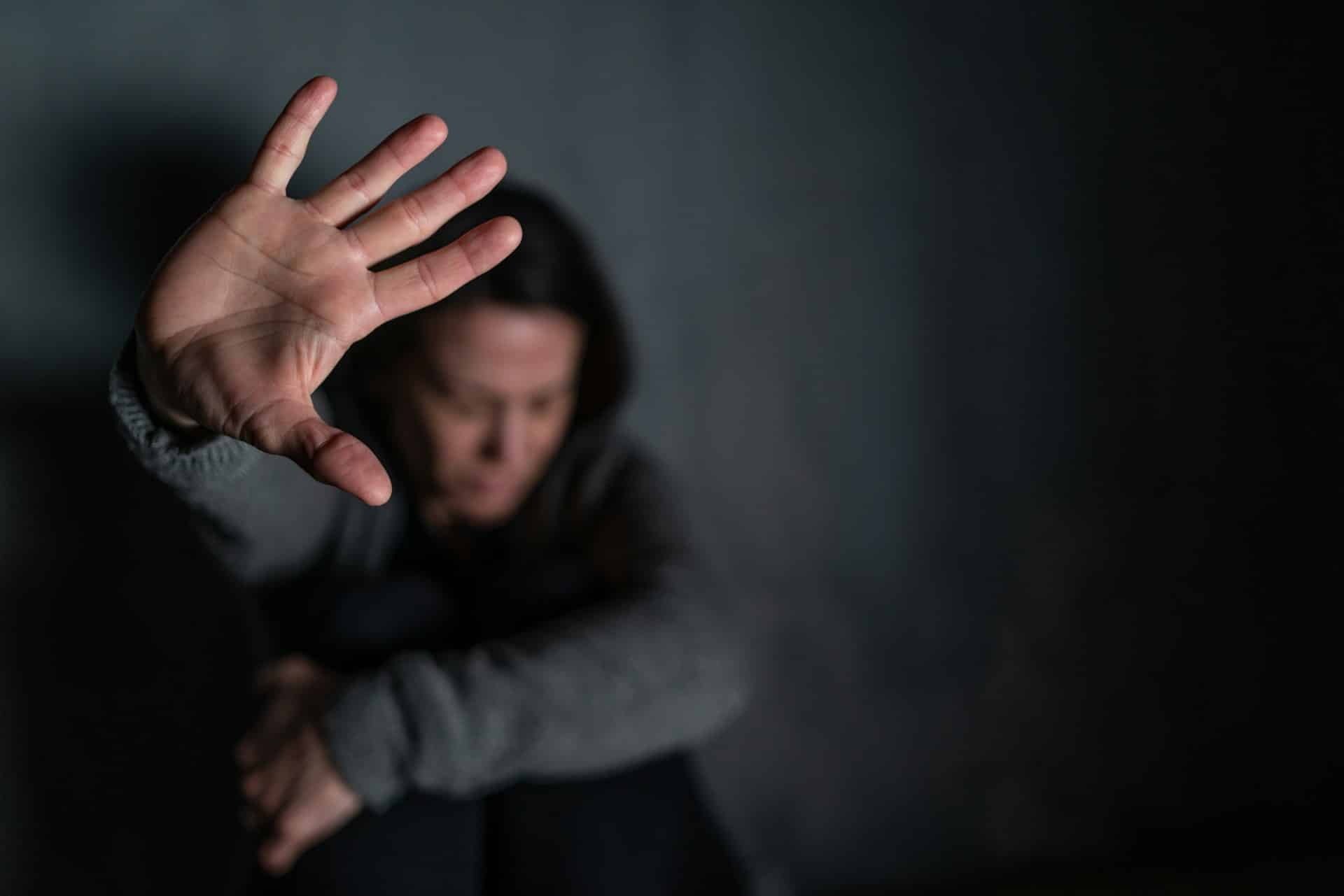 A visibly distressed woman is crouched on the floor against a wall with one arm wrapped around her knees, drawn to her chest, while the other is raised with her hand up and palm out, representing family violence and emergency protection orders in Alberta.