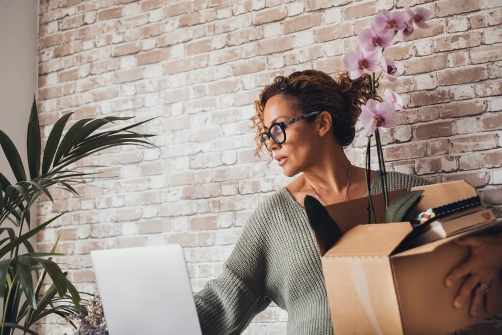 A terminated woman carries a box with her workplace belongs in one hand while she reaches for her laptop with the other as she clears out her office, representing reasonable notice calculations.