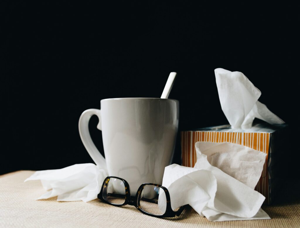 A mug with a spoon inside sits next to a box of tissues and crumpled up tissues, and glasses on a table, representing job-protected sick leave.