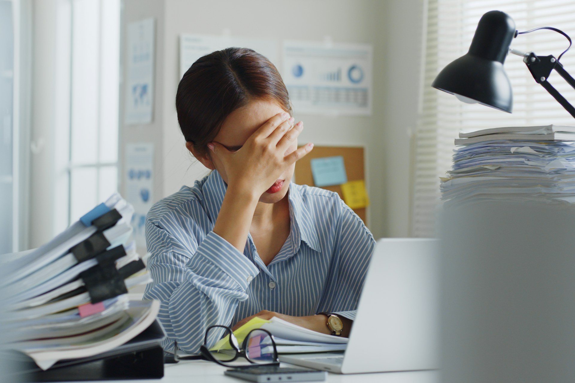 A tired employee rests her forehead in her hand with her laptop open in front of her and glasses on the desk next to it, surrounded by stacks of paper, representing quiet hiring risks in Alberta.