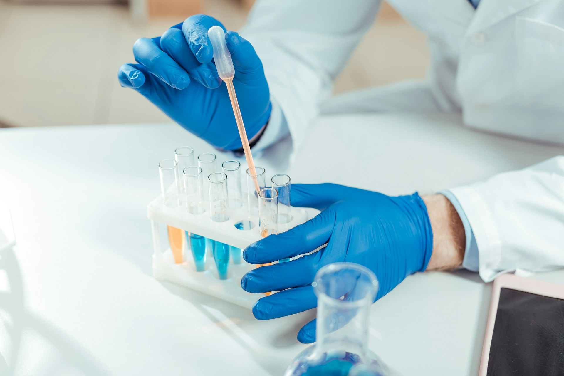 A medical professional, shown from the shoulders down, wearing blue latex gloves test blue and orange liquid in test tubes, representing drug & alcohol testing policies.